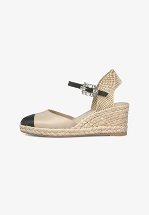 Alpargata cuña beige con puntera negra, tira de talón tejida y tira de tobillo negra con una hebilla decorativa de pedrería.