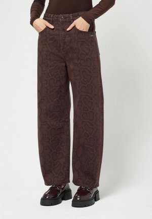 Personne portant un pantalon marron à jambes larges avec imprimé serpent, les mains dans les poches et des mocassins sombres à semelles épaisses.