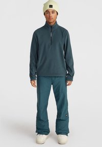 Tumedast türkiissinises fleecist pullover, millel on neljandiku varrukas ja logo; koos lahtiste türkiissiniste pükstega ja kreemivärvi lumepükstega.
