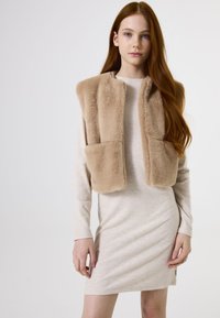 Jonge vrouw met lang roestbruin haar, gekleed in een beige ribbeljurk en een tan nepbont vest, tegen een effen witte achtergrond.