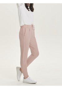 Kvinna iklädd ljusrosa slim-fit byxor med sidostreck och vita sneakers, stående med ett ben korsat över det andra.