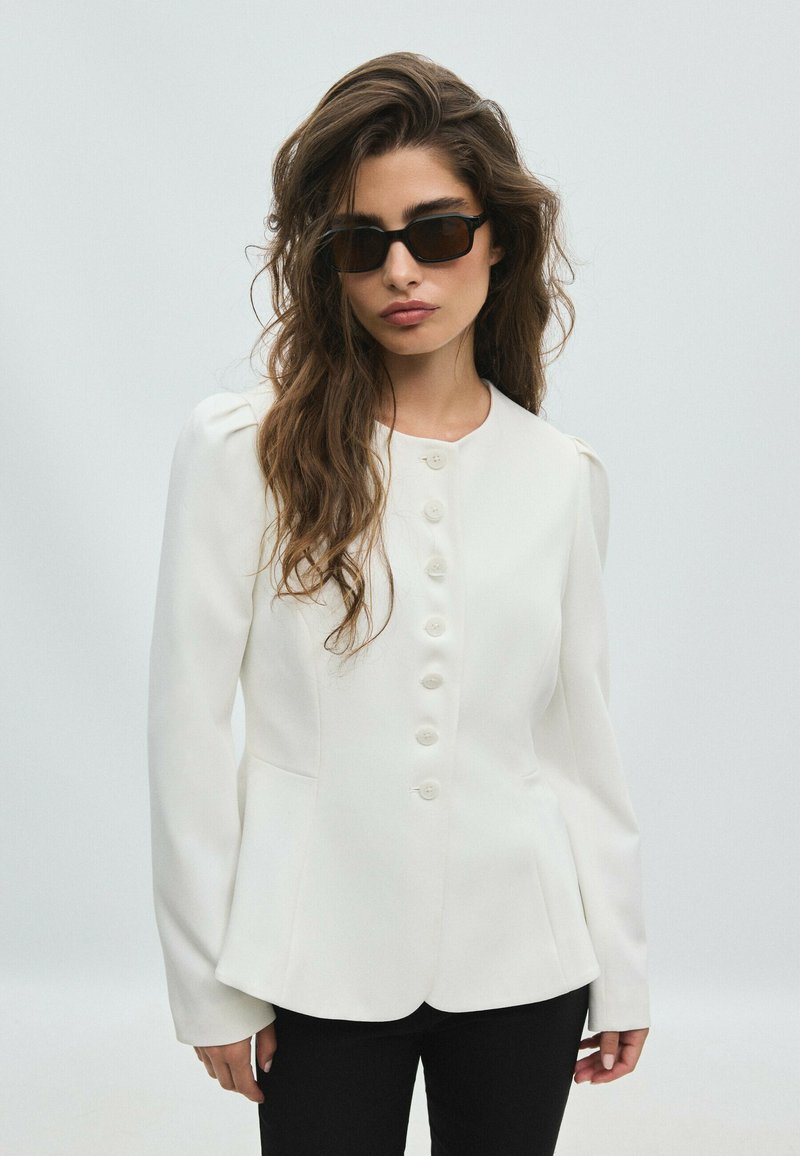 Femme aux longs cheveux ondulés portant des lunettes de soleil noires rectangulaires, une veste blanche boutonnée et un pantalon noir sur un fond uni.