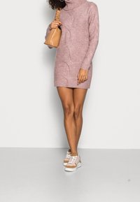 Robe à col roulé en tricot rose avec des détails en tricot à côtes. Associée à des baskets beiges avec des accents roses. La robe se termine au-dessus des genoux.