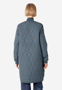 Abrigo largo acolchado en verde azulado oscuro, con un patrón de diamantes, cuello y puños de canalé. El diseño es funcional y minimalista.