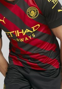 Camiseta de fútbol del Manchester City a rayas negras y rojas con el logo amarillo de Puma y el texto del patrocinador Etihad Airways, combinada con pantalones cortos negros.
