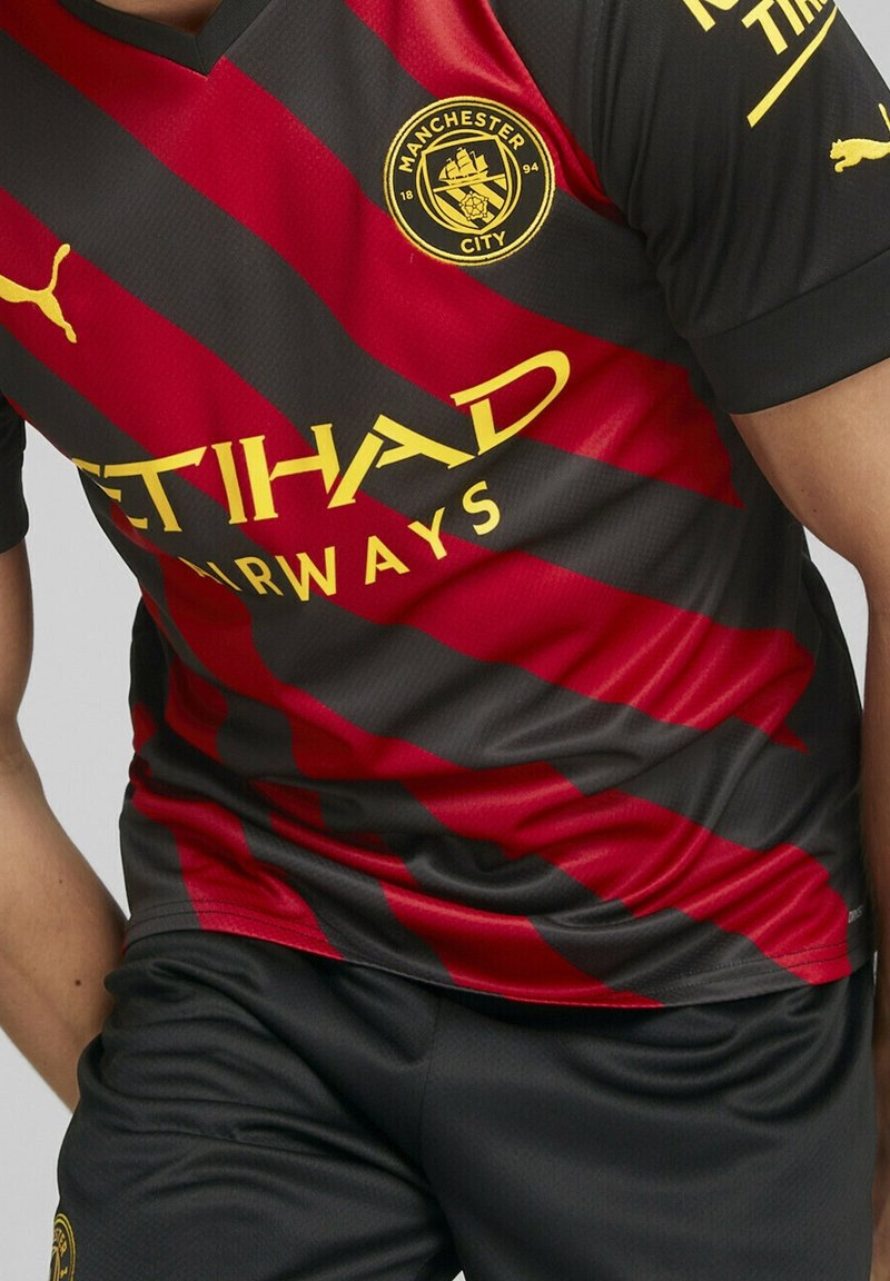Camiseta de fútbol del Manchester City a rayas negras y rojas con el logo amarillo de Puma y el texto del patrocinador Etihad Airways, combinada con pantalones cortos negros.