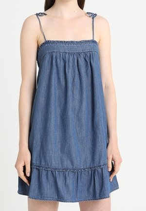 Frau trägt ärmelloses blaues Jeanskleid mit dünnen Trägern, die an den Schultern gebunden sind, und Rüschensaum vor weißem Hintergrund.