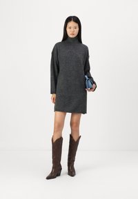 Grå strikket polo-kjole med lange ermer, midt-lår lengde. Modellen har på seg brune cowboy-støvler og holder en liten blå veske.