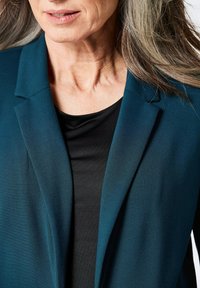 Teal blazer met een notch lapel, gecombineerd met een zwarte top. De blazer heeft een soepele textuur en een gestructureerd ontwerp, dat de halslijn accentueert.