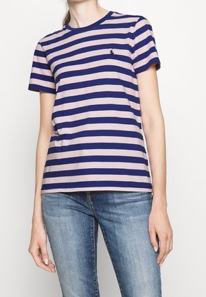 Kvinde iført en kortærmet t-shirt med horisontale striber i navy og lyserød samt blå jeans mod en ensfarvet hvid baggrund.