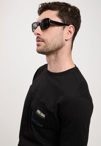 Czarna koszula z długim rękawem i kieszenią na piersi, z metką. Model nosi błyszczące czarne prostokątne okulary przeciwsłoneczne. Proste tło.