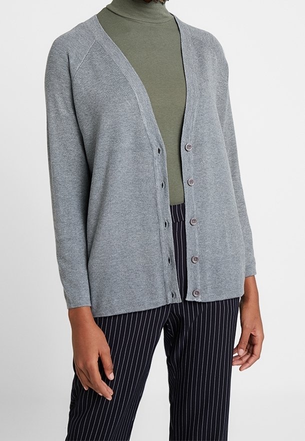 Femme portant un cardigan à boutons gris clair par-dessus un col roulé vert olive avec un pantalon noir à fines rayures, debout devant un fond uni.