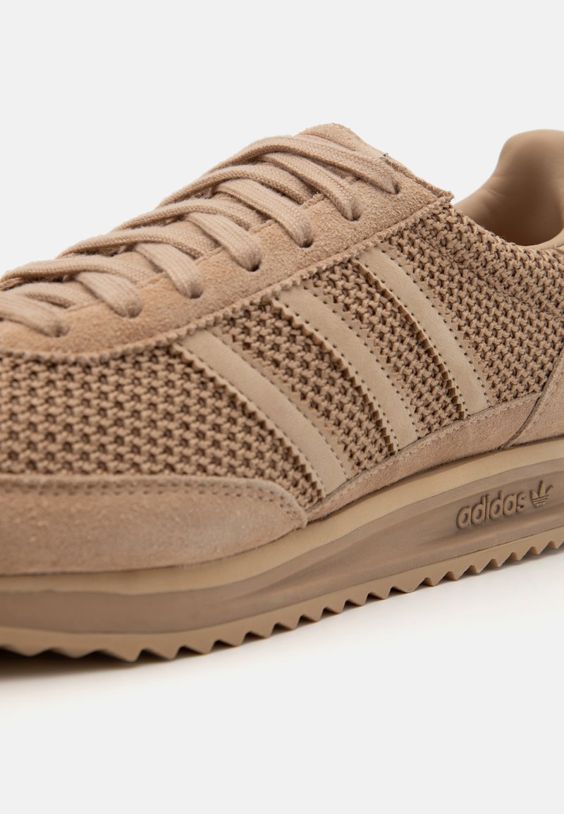 Zapatilla deportiva beige con parte superior de malla, tres franjas laterales texturizadas, suela de goma con un patrón en zigzag y un collar acolchado.