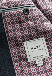 Maßgeschneiderte Jacke mit einer dunkelblauen Textur, ausgestattet mit einem fächerförmigen Futter in Bordeauxrot, Grau und Weiß, und einem Etikett, das "NEXT Tailoring, Regular Fit" besagt.