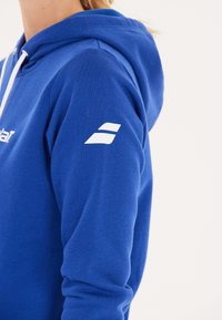 Blå hoodie lavet af bomuldsblanding, med en snoretrækshætte og hvid logo detalje på ærmet. Glat tekstur og afslappet pasform.