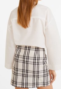 Chemise blanche à col avec de longues manches, associée à une jupe mini ajustée à carreaux dans des tons beige, noir et gris ; texture de tissu lisse.