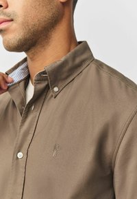 Bruine overhemd met knopen en een kraag, met een subtiel geborduurd ontwerp en lichtblauwe gestreepte accenten zichtbaar aan de binnenkant van de kraag.
