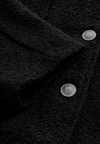 Tessuto nero testurizzato con superficie pelosa; presenta due bottoni d'argento con incisioni decorative; design elegante e moderno.