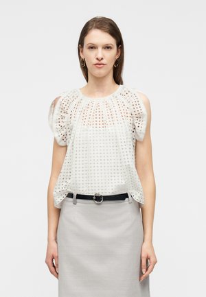 Femme portant un chemisier blanc en broderie anglaise avec découpe aux épaules dénudées, associé à une jupe gris clair et une ceinture noire à boucle ronde.