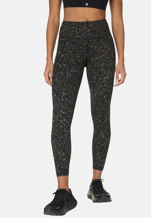 Vrouw draagt zwarte hoge taille legging met meerkleurige spikkels en zwarte sportschoenen, staand tegen een effen achtergrond.