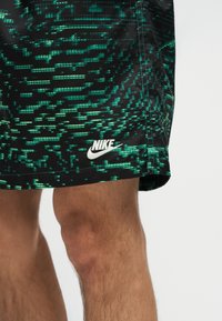 Μερική εμφάνιση ποδιών που φορούν μαύρα και πράσινα αθλητικά σορτς Nike με σχέδιο, με λευκό λογότυπο Nike κοντά στο τελείωμα.