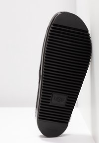 Semelle de chaussure noire avec de profondes rainures horizontales et le logo UGG embossé près du centre, reposant contre une surface blanche.