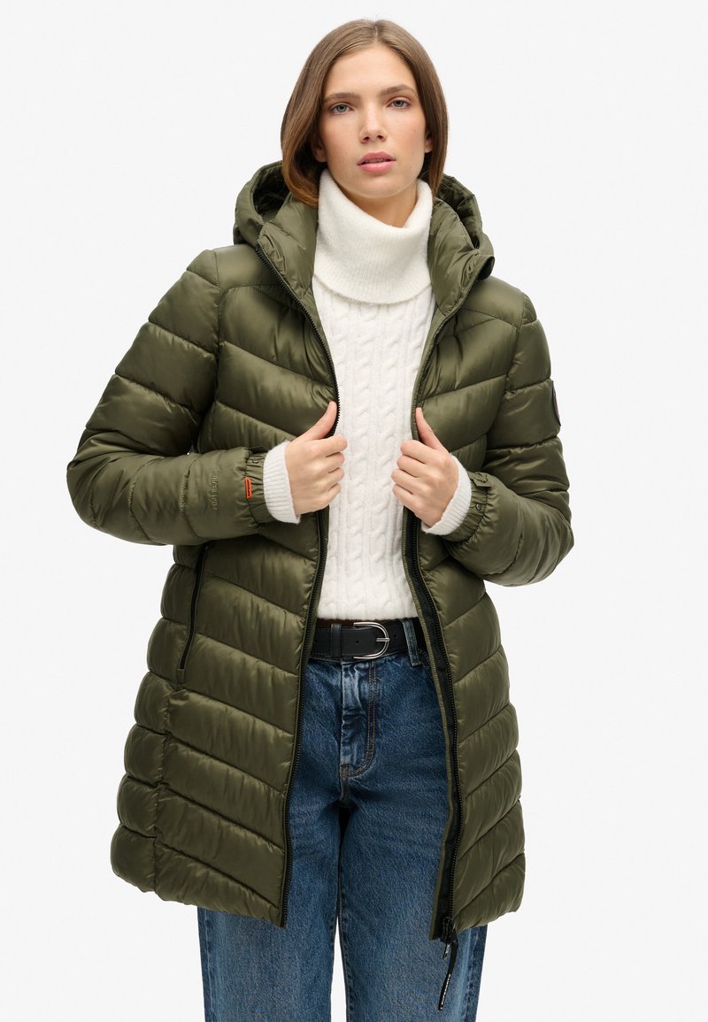 Abrigo tipo puffer verde oliva con capucha, con diseño acolchado, cremallera frontal y puños de canalé, combinado con un suéter blanco de punto cableado y jeans.