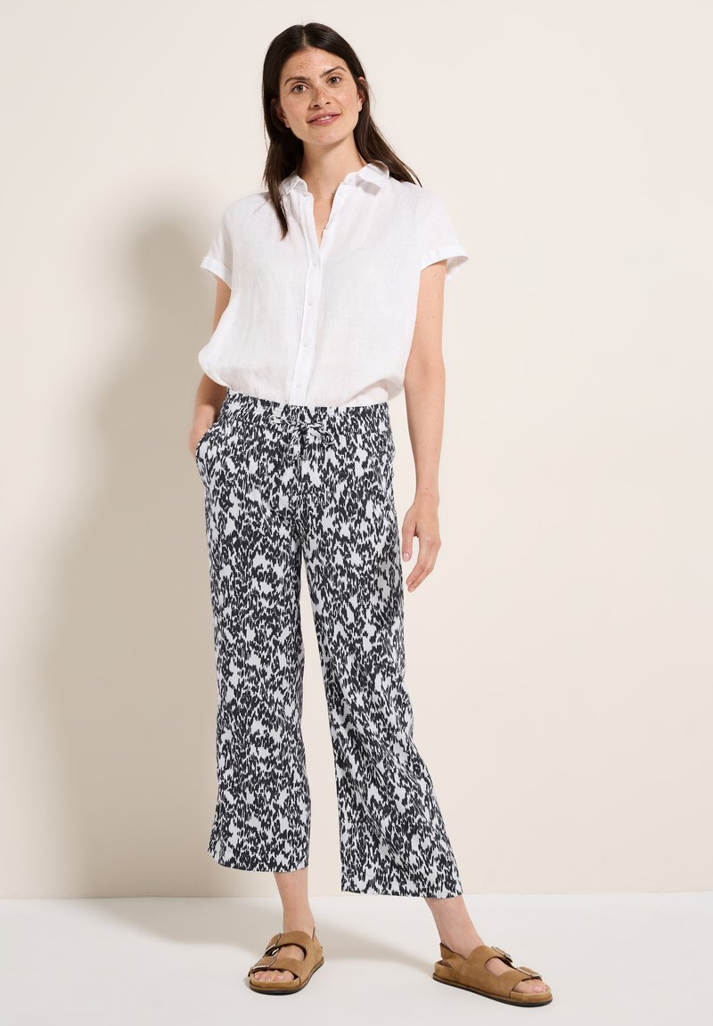 Witte linnen kortarmige shirt, met een patroon in zwart en wit brede broek met aansnoerbare taille, en bruine platte sandalen.