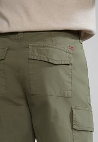 Plano cercano de pantalones cargo verde olivo con bolsillos con solapa y un pequeño parche de la bandera noruega en el bolsillo trasero, combinados con una blusa beige.