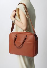Bolso de cuero marrón con asas dobles en la parte superior y una correa de hombro desmontable. Textura suave, forma rectangular y un sutil detalle del logo en la parte frontal.