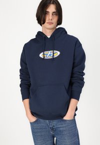 Haut en coton bleu marine avec une poche avant, capuche à cordon et un logo graphique circulaire jaune et bleu sur la poitrine.