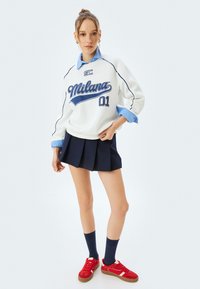 Sudadera blanca con la inscripción azul "Milana" sobre una camisa de color azul, combinada con una falda plisada navy y deportivas rojas.