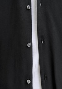 Vêtement noir boutonné partiellement ouvert pour révéler un tissu blanc texturé en dessous, montrant trois boutons et trois boutonnières sur le tissu noir.