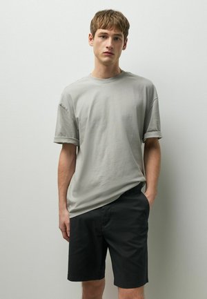 Noor mees lühikeste pruunide juustega, seljas lahtine helehall T-särk ja mustad lühikesed püksid, seisab lihtsa heleda seina taustal.