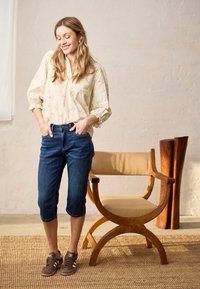 Femme souriante en blouse légère à fleurs, pantalon en denim court et baskets, debout les mains dans les poches à côté d'une chaise en bois dans une pièce lumineuse.