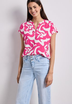 Mujer sonriendo, con una blusa de patrón abstracto en rosa y blanco y unos vaqueros azul claro, de pie frente a un fondo liso y claro.