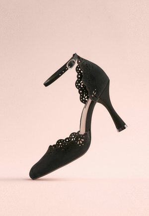 Sort højhælet sko med spids tå, bølgede udskæringer, justerbar ankelrem og struktureret ruskindsoverflade.