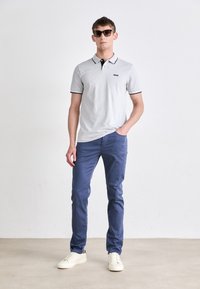 Polo de color gris claro con cuello y detalles en negro, combinado con chinos azules y zapatillas crema. Modelo con gafas de sol oscuras.