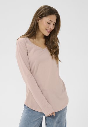 Mladá žena s dlhými vlasmi, ktorá nosí zlaté kruhové náušnice, ružové dlhé tričko s výstrihom do V a modré džínsy, usmieva sa a pozerá dole.