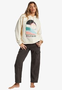 Hoodie crema con design grafico che presenta un tramonto e una spiaggia, abbinato a pantaloni cargo scuri. Il hoodie ha corde di regolazione e polsini a coste.