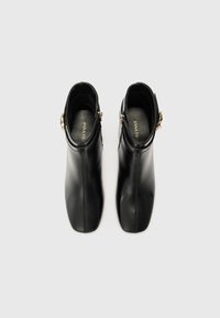 Bottes noires en cuir à la cheville avec une finition lisse, un bout pointu et des éléments décoratifs en métal doré sur les côtés.