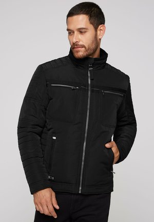 Schwarze gepolsterte Jacke mit hohem Kragen, Reißverschluss, Brust- und Seitentaschen, gerippten Schultereinsätzen und strukturierten Stoffpaneelen.