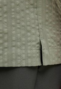 Olive green textile with a textured pattern, featuring horizontal ribbing and a side split at the hem. Smooth fabric and tailored fit.