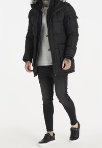 Svart puffjacka med foder av päls i huvan, dragkedjefickor, framslående knappar, bärs över en grå skjorta och kombineras med mörka jeans och svarta sneakers.