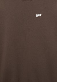 Rjava bombažna majica z mehko teksturo, z majhnim belim logotipom na zgornji levi strani, z okroglim izrezom in kratkimi rokavi.