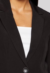 Czarna marynarka o dopasowanym kroju z wyciętymi klapami, jednogniazdowym zapięciem i miękką teksturą tkaniny. Noszona na białej, dopasowanej bluzce.