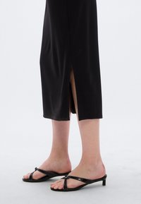 Μαύρο midi φόρεμα με πλαϊνή σχισμή, κατασκευασμένο από λείο υλικό. Φορε μένο με μαύρα σανδάλια με λουράκια, χαμηλό τακούνι και μεταλλική αγκράφα.