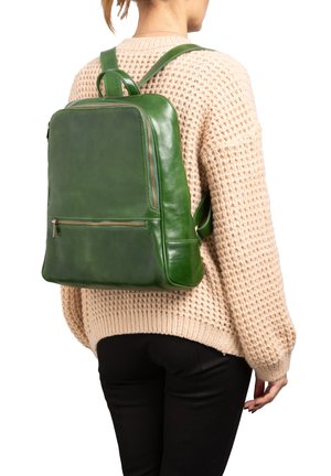 Groene leren rugzak met een rechthoekige vorm, ritssluiting aan de voorkant en verstelbare schouderbanden, gedragen over een gebreide beige sweater.