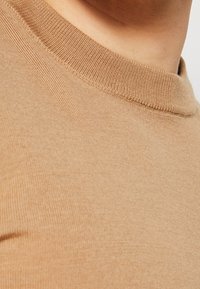 Gros plan sur un pull beige en maille à col rond porté par une personne, montrant la texture et les détails du tissu sur l’épaule et la zone du cou.