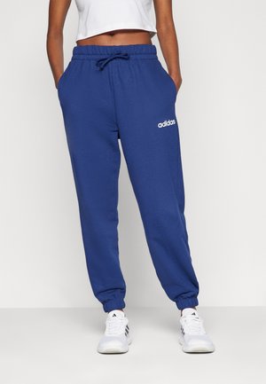 Personne portant un pantalon de survêtement Adidas bleu avec des poignets élastiques et un crop top blanc, debout les mains dans les poches avec des baskets blanches Adidas.